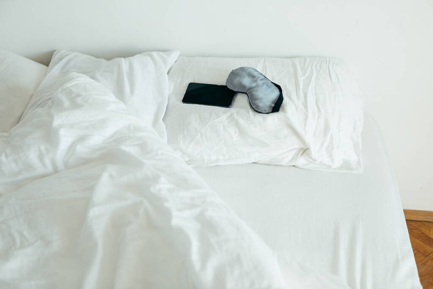 Beyaz çarşaflı, güneşli yatak odalı, telefonlu uyku maskesi. - Fotoğraf, Görsel