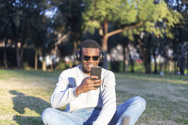 Fashionable male with dark skin and sunglasses sitting outdoors checking his news feed or messaging via social networks using cell phone - Foto, Imagen