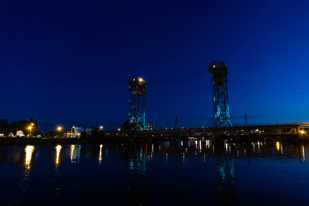 duży żelazny most z dwiema wysokimi wieżami nad rzeką w świetle latarni w nocy - Zdjęcie, obraz