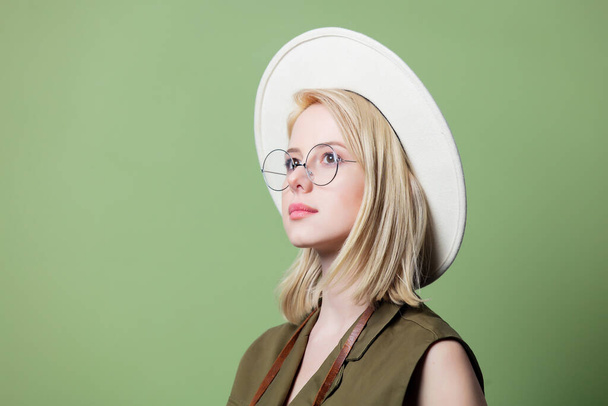 Yeşil arka planda gözlüklü, şapkalı ve elbiseli zarif sarışın kadın. - Fotoğraf, Görsel