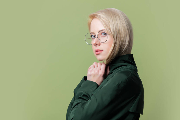 Styl blondýny žena v plášti a kulaté brýle na zeleném pozadí  - Fotografie, Obrázek