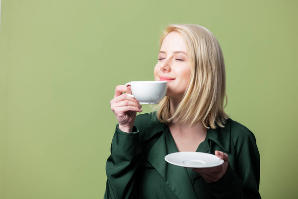 Blonde woman in cloak with cup of coffee on green background  - Valokuva, kuva