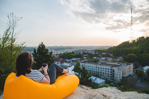 mujer sentada en colchón inflable amarillo bebiendo café disfrutando del atardecer sobre la ciudad. espacio de copia
 - Foto, imagen