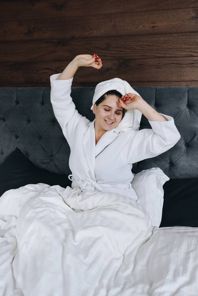 Молодая кавказская женщина растягивается в постели в спальне с полотенцем на голове
 - Фото, изображение