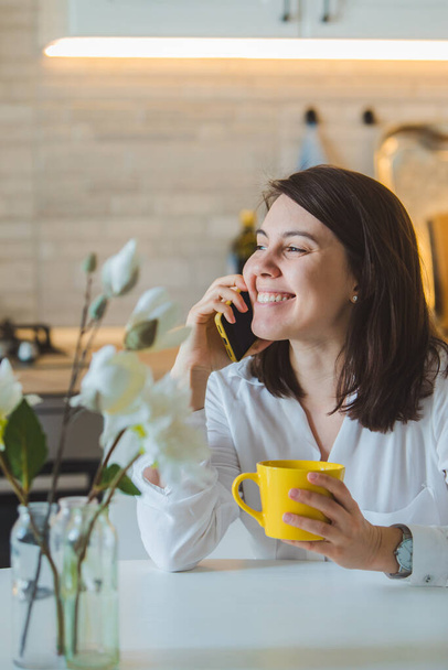 mulher bebendo chá de caneca amarela na cozinha falando ao telefone
 - Foto, Imagem