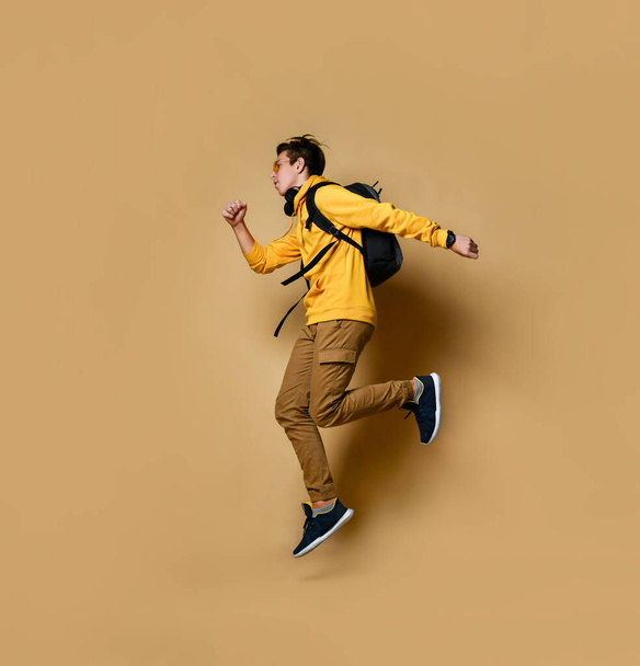 Young teen boy in comfortable clothing, sneakers and backpack jumping over yellow wall background - Фото, зображення