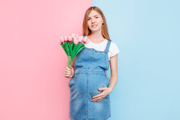 Happy beautiful pregnant woman with a bouquet of tulips, posing standing on a pink and blue background. The concept of mother's Day - Valokuva, kuva