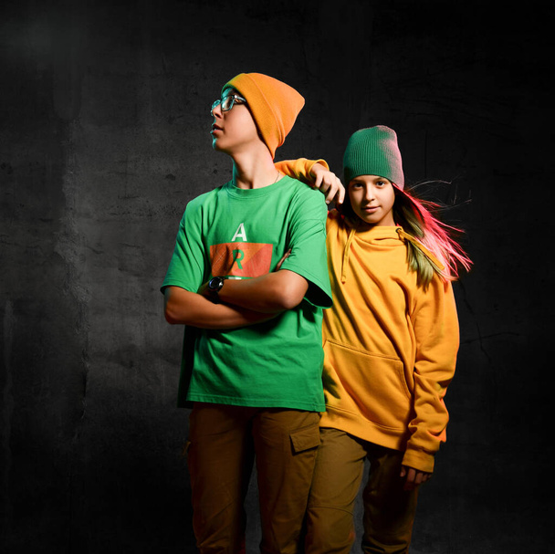 Young teens boy and girl in green and yellow stylish comfortable casual clothing and hats standing over dark background - Valokuva, kuva