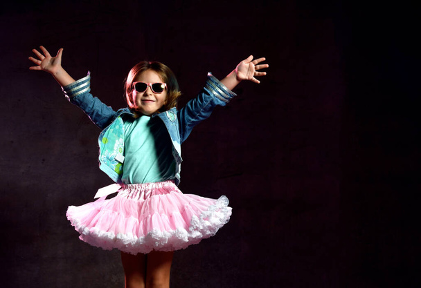 Little girl in blue t-shirt, denim jacket, poofy skirt, sneakers, sunglasses. Bouncing up against black background. Full length - Φωτογραφία, εικόνα