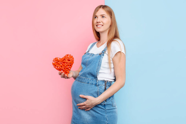 Happy beautiful pregnant woman holding a red heart near her belly, standing on a pink and blue background - Zdjęcie, obraz