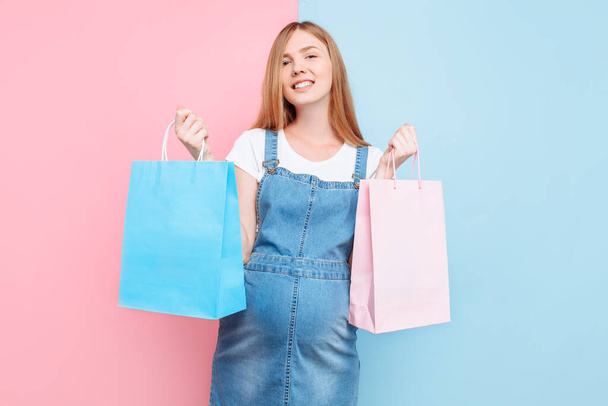 A beautiful pregnant young woman holds a pink and blue shopping bag, on an isolated pink and blue background. Determine the gender of the child: twins, girl or boy. - Fotoğraf, Görsel