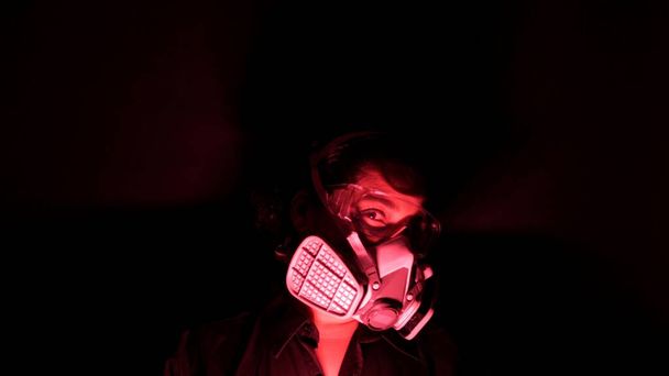 Woman in the heavy duty biohazard medical mask in the darkness and red light. Personal forced home quarantine isolation from Coronavirus or covid 19 concept. Apocalypses and depression mood.  - Foto, imagen