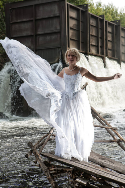 jonge blonde vrouw in een witte bruiloft jurk in de buurt van de waterval - Foto, afbeelding