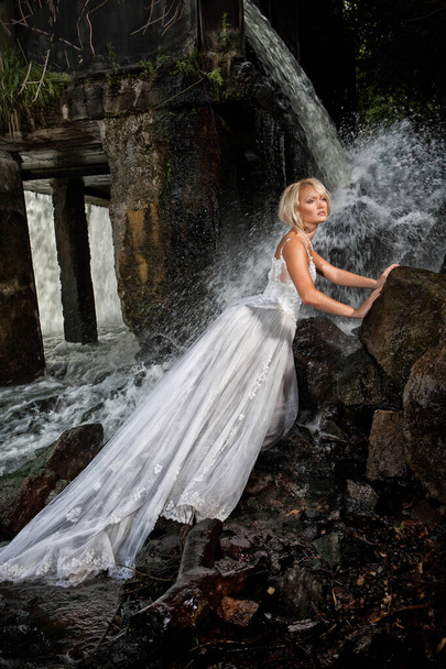 junge blonde Frau in einem weißen Hochzeitskleid in der Nähe des Wasserfalls - Foto, Bild