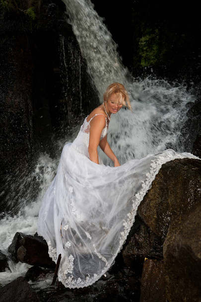 Young blonde woman in a white wedding dress near the waterfall - Foto, afbeelding