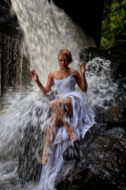 Young blonde woman in a white wedding dress near the waterfall - Фото, изображение