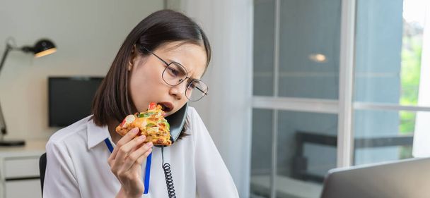 Jovem empresária asiática fazendo lanche de pizza enquanto conversa telefone com clientes devido ao trabalho até tarde
. - Foto, Imagem