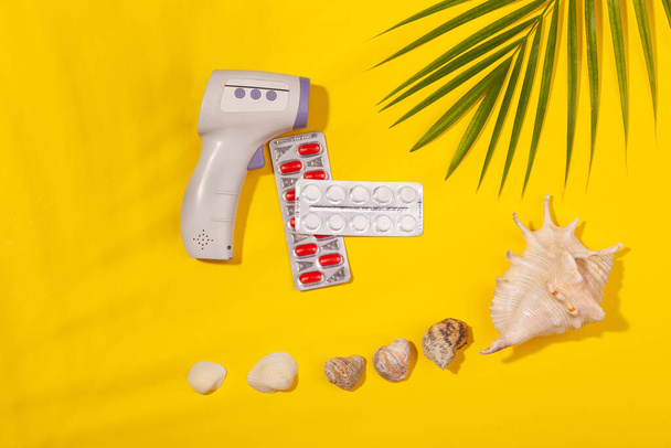 Top view on infrared thermometer, white and red pills in blister pack, seashells, palm leaf on yellow background. Concept of beach holiday, sea tour, warm sunny summer. Concept of preparing for - Foto, afbeelding