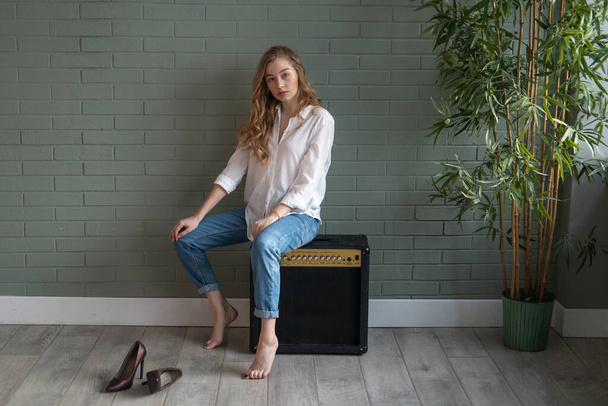 Young beautiful blond woman wearing white shirt and blue jeans sitting on subwoofer with bare feet next to green plant. Girl sitting in a daylight from the window. Copy space - Foto, Imagem