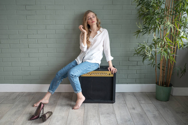 Young beautiful blond woman wearing white shirt and blue jeans sitting on subwoofer with bare feet next to green plant. Girl sitting in a daylight from the window. Copy space - 写真・画像