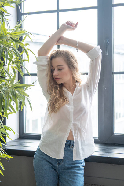 Young beutiful blond woman wearing white shirt and blue jeans standing next to window. Morning portrait of a girl with window and green plants on the background - Photo, image