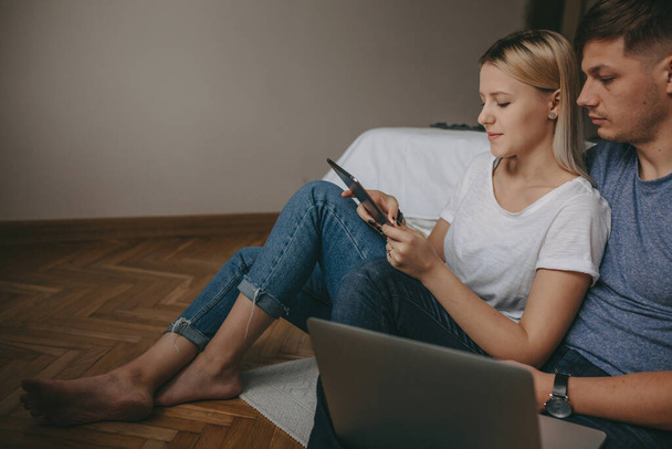 Casal caucasiano sentado no chão e usando um laptop e tablet enquanto procura algo
 - Foto, Imagem