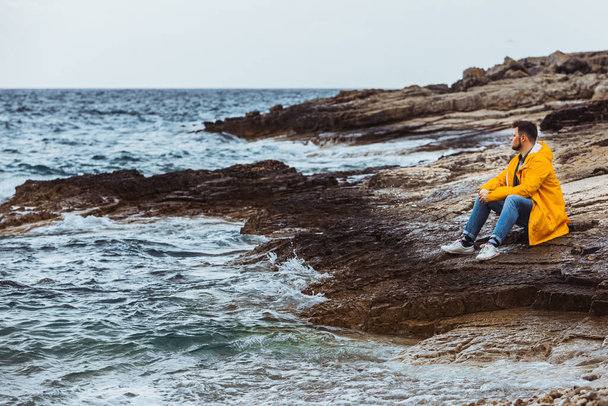uomo seduto in impermeabile giallo sulla spiaggia rocciosa guardando il tempo tempestoso. copia spazio. - Foto, immagini