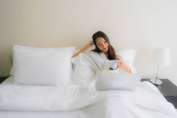 Портрет красивые молодые азиатские женщины с чашкой кофе и ноутбуком на кровати в интерьере спальни - Фото, изображение