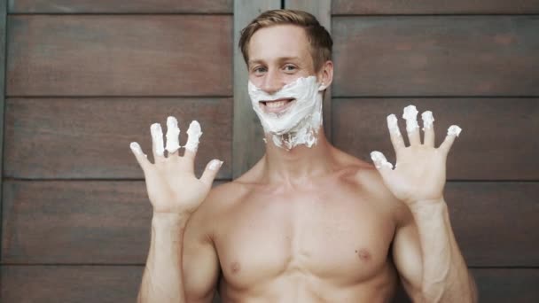 Portrait of happy man puts shaving foam on face, laughs and shows ok sign - Filmati, video
