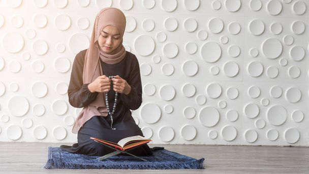 Retrato de una hermosa joven musulmana sentada en la alfombra de oración usando cuentas de rosario mientras lee el quran con un patrón redondo fondo blanco
 - Foto, Imagen