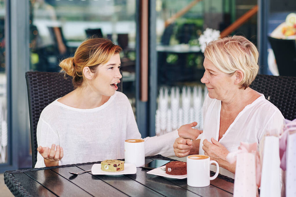 Kaunis nuori nainen kertoo järkyttäviä uutisia ja juoruja äidilleen, kun he juovat kahvia ja jälkiruokia kahvilassa.
 - Valokuva, kuva