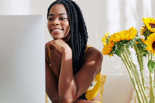 Gülümseyen yaratıcı genç siyahi kadın masasında bilgisayarla çalışıyor. - Fotoğraf, Görsel