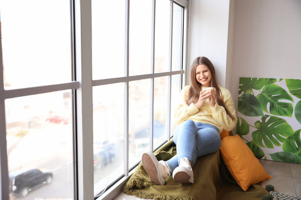 Όμορφη νεαρή γυναίκα με φλιτζάνι τσάι κάθεται στο περβάζι παράθυρο στο σπίτι - Φωτογραφία, εικόνα