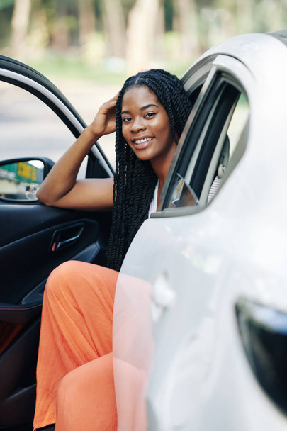 Atraktivní usmívající se mladá černoška sedí ve svém novém autě a dívá se do kamery - Fotografie, Obrázek