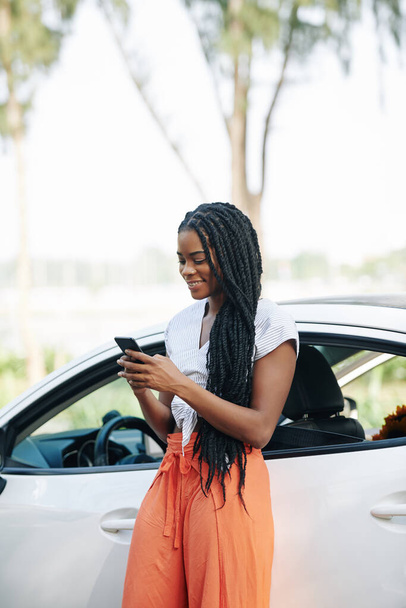 Θετική νεαρή μαύρη γυναίκα στέκεται στο αυτοκίνητό της και ελέγχει ειδοποιήσεις στο smartphone της - Φωτογραφία, εικόνα