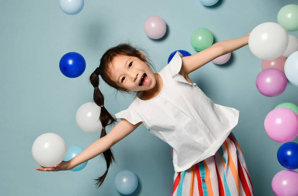 Joyful asian kid girl with long black plait is having fun surrounded by colorful air balloons, spread hands - Foto, Imagem