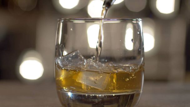Primer plano verter whisky en vaso de beber con hielo cubos. Bar contador en el fondo de las bombillas borrosas
. - Foto, Imagen