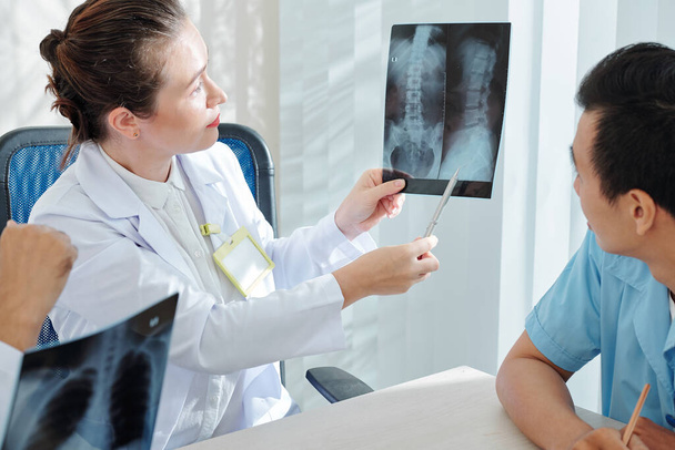 Mujer joven seria mostrando radiografía de columna curvada a compañeros de trabajo y discutiendo caso difícil
 - Foto, imagen