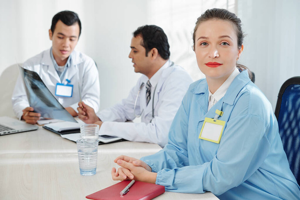 Впевнена позитивна жінка загальної практики сидить за великим столом у залі для лікарів
 - Фото, зображення