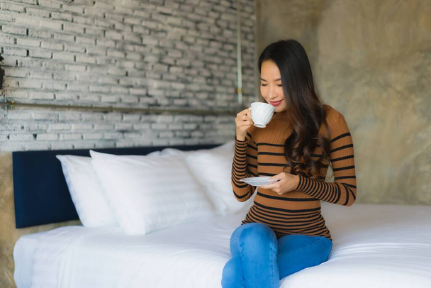 Νεαρή Ασιάτισσα με φλιτζάνι καφέ στο κρεβάτι στο εσωτερικό της κρεβατοκάμαρας - Φωτογραφία, εικόνα