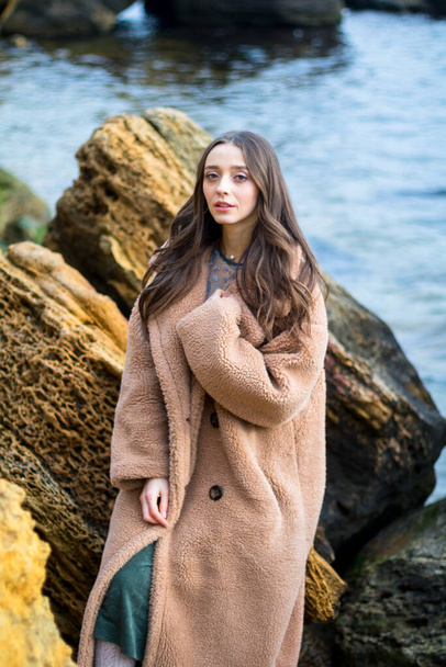 young sensual girl in brown fur coat is happy to be near the big yellow stones and sea water - Photo, Image