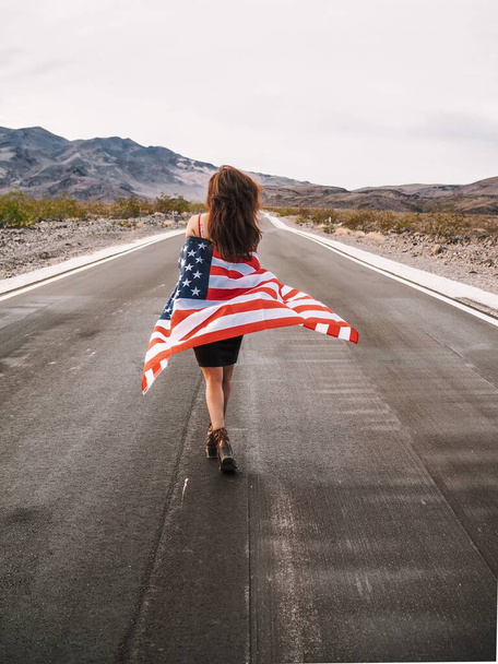 Brunette meisje met lang haar in een rok loopt over een lege weg in de vallei van de dood zwaaiend met de Amerikaanse vlag - Foto, afbeelding