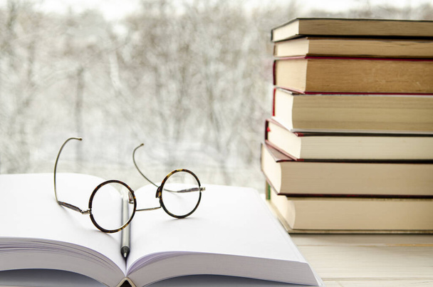 Круглые очки для чтения лежат на открытой книге с белыми страницами рядом со стопкой разноцветных книг на деревянном подоконнике - Фото, изображение