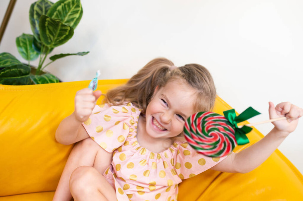 portrait of a happy girl with a candy. The child sits on the yellow sofa and laughs, indoor - Photo, Image