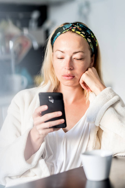 Beautiful caucasian woman at home, feeling comfortable wearing white bathrobe, taking some time to herself, drinking morning coffee and reading news on mobile phone device in the morning - Valokuva, kuva