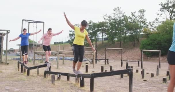 Side view of a happy multi-ethnic group of women enjoying exercising at boot camp together, balancing and walking along a wooden beam in slow motion - Filmati, video