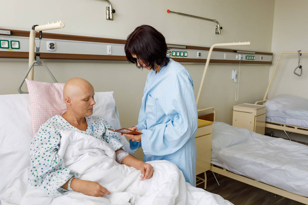 Woman without hair after chemotherapy patient lying at hospital bed feeling sad and depressed worried. A friend came to please her, showing photos on her mobile phone. - Zdjęcie, obraz