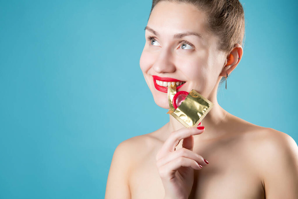 Girl playfully holds the edge of the wrapper of a red condom with her teeth - Фото, изображение
