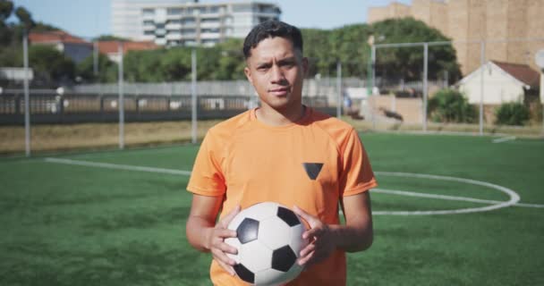 Portrait of a mixed race male football player wearing sportswear, looking at camera, holding a football, standing on a sports field, in slow motion - Materiaali, video