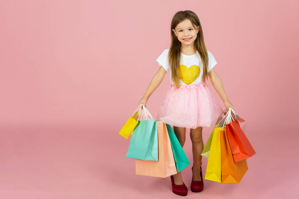 Beautiful cute little girl with long blonde hair, tulle skirt standing in mothers big shoes with colorful packages isolated on pink background. Expressing true positive emotions to camera. - Photo, Image